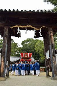 濃い茶色の木材で造られた立派な門の奥で、鮮やかな紅色の神輿を青い法被を着た大人数の大人の人が担いでいる様子の写真