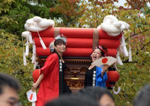 左側に赤い法被をきた女性と、右側に青い法被を着た女性が神輿の上に立ち、アップで映る様子の写真