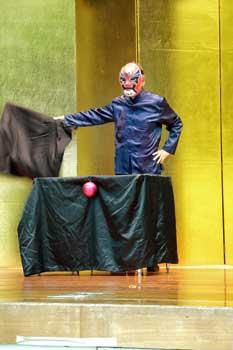 ステージ上で仮面をつけた男性が右手に茶色の布状のものを持っている写真