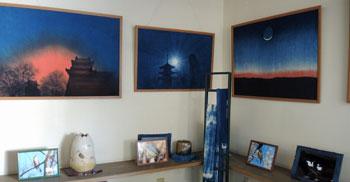 藍染めで描かれた景色の大きな作品が壁に3点かけられ、その下の台に他の写真作品が展示されている写真