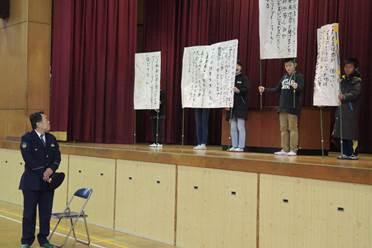 ステージ下のお巡りさんとステージで歌の歌詞が書かれた紙を拡げている小学生の写真
