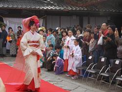 観覧者に囲まれた絨毯の上を歩く白い着物に赤いベールをまとった女性の写真