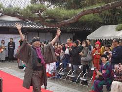 観覧者に囲まれた赤い絨毯の上を歩く茶色の着物を着た男性が両手を挙げて笑顔で歩いている写真
