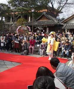 観覧者に囲まれた赤い絨毯のうえで黄色い着物を着たが両腕を広げてポーズをとっている写真