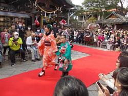 観覧者に囲まれた赤い絨毯を歩くオレンジの着物と緑の着物を着た二人の女の子の写真