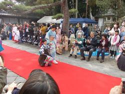 観覧者に囲まれた赤い絨毯を歩く水色の着物の女性と袴をはいた男の子の写真