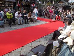 石畳の境内に敷かれた赤い絨毯の両脇にいる観覧者の写真