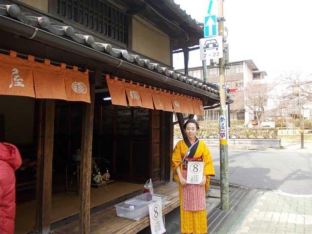 短い暖簾がかかった日本家屋の前に黄色の着物を着た女性が立っている写真