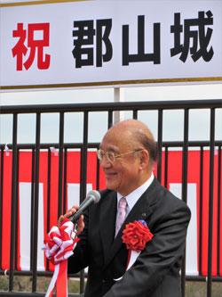 紅白の幕を背に胸に赤い花を付けたスーツの男性がスピーチをしている写真