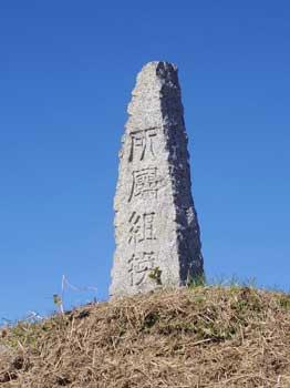 雲一つ無い抜けるような濃い青空の下、芝の丘の上に立つ「所属組換」と彫られた縦長の石塔状の石碑の写真