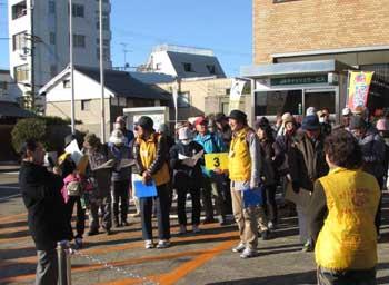 駅前の広場で黄色いベストを着用したガイドの3人の男女とその後ろに控える大勢の参加者を前に、話をしているメガネを付けてコートを着た男性の写真