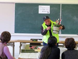 蛍光カラーチョッキを着た男性講師がミニ門松の完成見本を公開している様子の写真