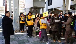 駅での集会で、 3人の黄色いチョッキを着た人が案内をしている写真
