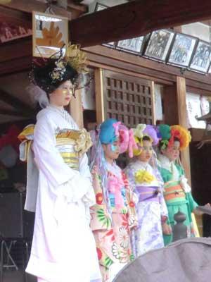 神社の拝殿の中、金色の煌びやかな髪飾りを付けた白い着物に金の帯を締めた女性と、その奥にそれぞれピンク色、紫色、緑色の着物を着てカラフルな髪飾りを付けた3人の女性が並んでいる写真