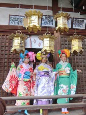 神社の拝殿の軒下、上から吊された金色に光る5つの飾り付きの台座の下に、左側にピンク色の着物を着た女性、真ん中に紫色の着物を着た女性、右側に緑色の着物を着た女性が3人並んでいる写真