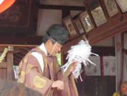 神社の拝殿の中で、黒い烏帽子を被り茶色い神主の装束を着た男性が、手に祭具を持ちながら一礼している写真