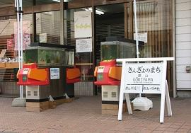 きんぎょのまちと書かれた駅名表示板とその隣に置かれている金魚の水槽を載せた2つの赤い改札機の写真