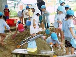 砂山で水を使って遊ぶ空色の帽子を被った保育園児たちの写真