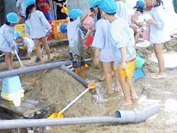 空色の帽子を被った保育園児たちが役割分担しながら砂場で水を使って遊んでいる写真
