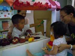 おもちゃを置いた手作りのカウンターを挟んで対面する小さい二人の子どもと後ろから支える二人の女性の写真
