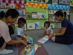 本棚の前で床に座り絵本を読み聞かせる二組の親子の写真