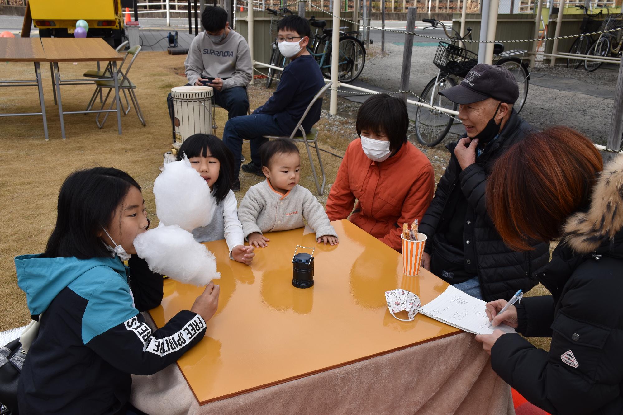 屋外の会場に置かれたコタツで綿菓子を食べる子供たちと祖父母