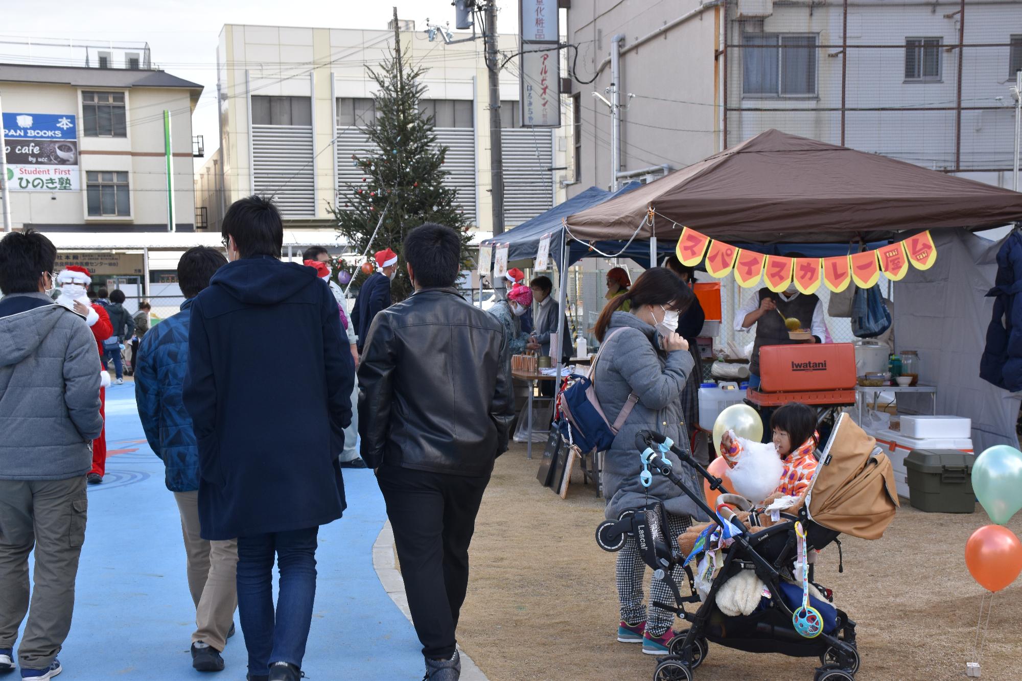 会場を訪れたコートを着た男性客4人とベビーカーに乗って綿菓子を食べる女の子とお母さん、テントを張って出店しているカレー屋