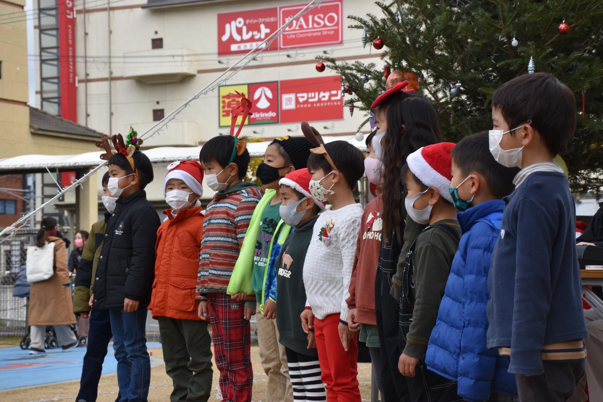 ツリーの前で、サンタの帽子やトナカイの角を頭に付けてクリスマスソングを歌う幼稚園児たち12人