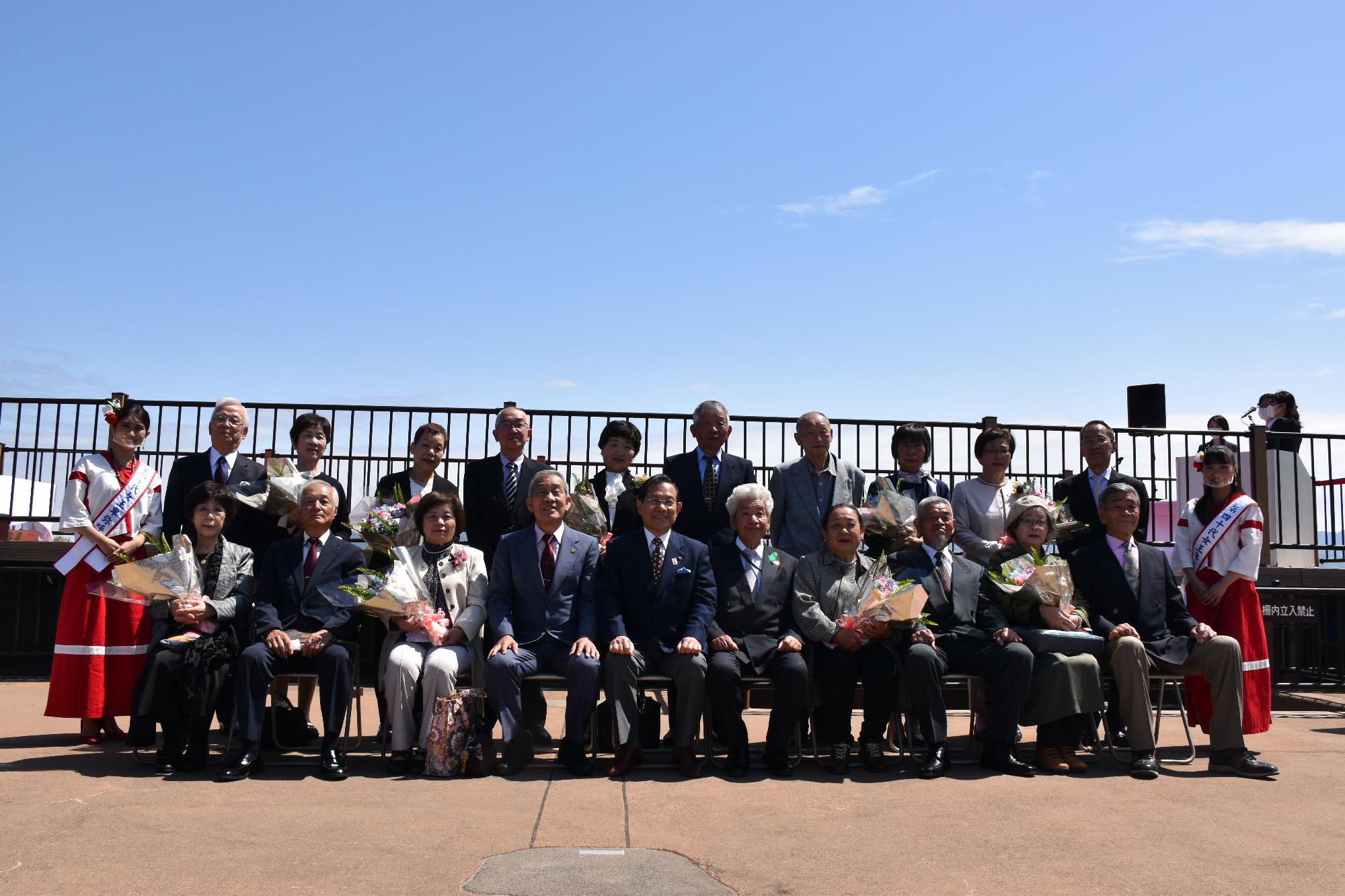 天守台展望施設で椅子に並んで座り記念撮影をする金婚式参加者夫婦9組と市長、女王卑弥呼の二人