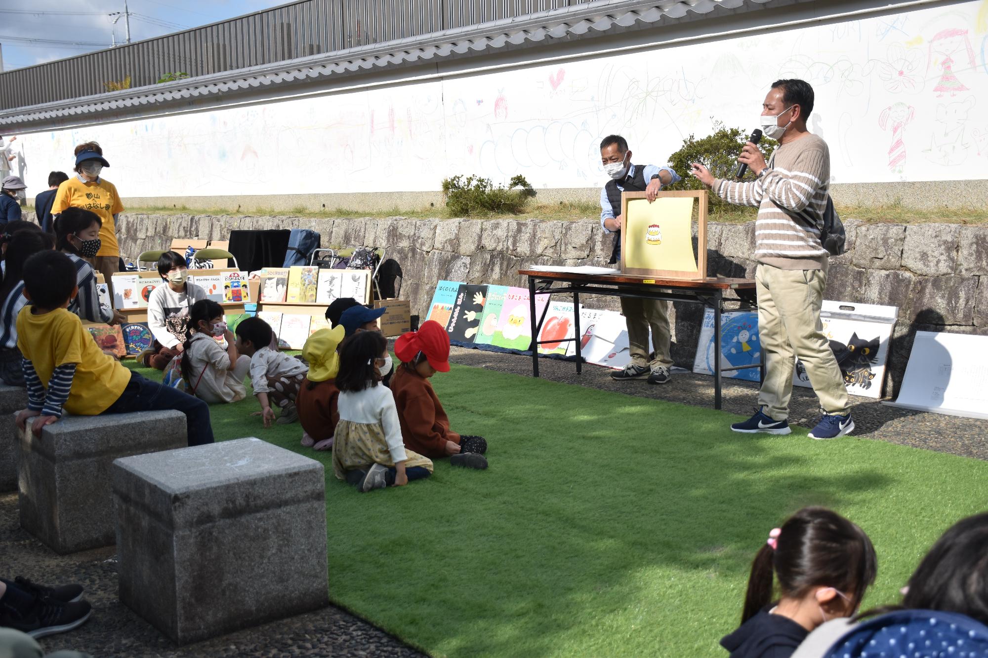 ベージュと白のストライプのシャツを着た紙芝居をする男性と前に座って聞いている子どもたち