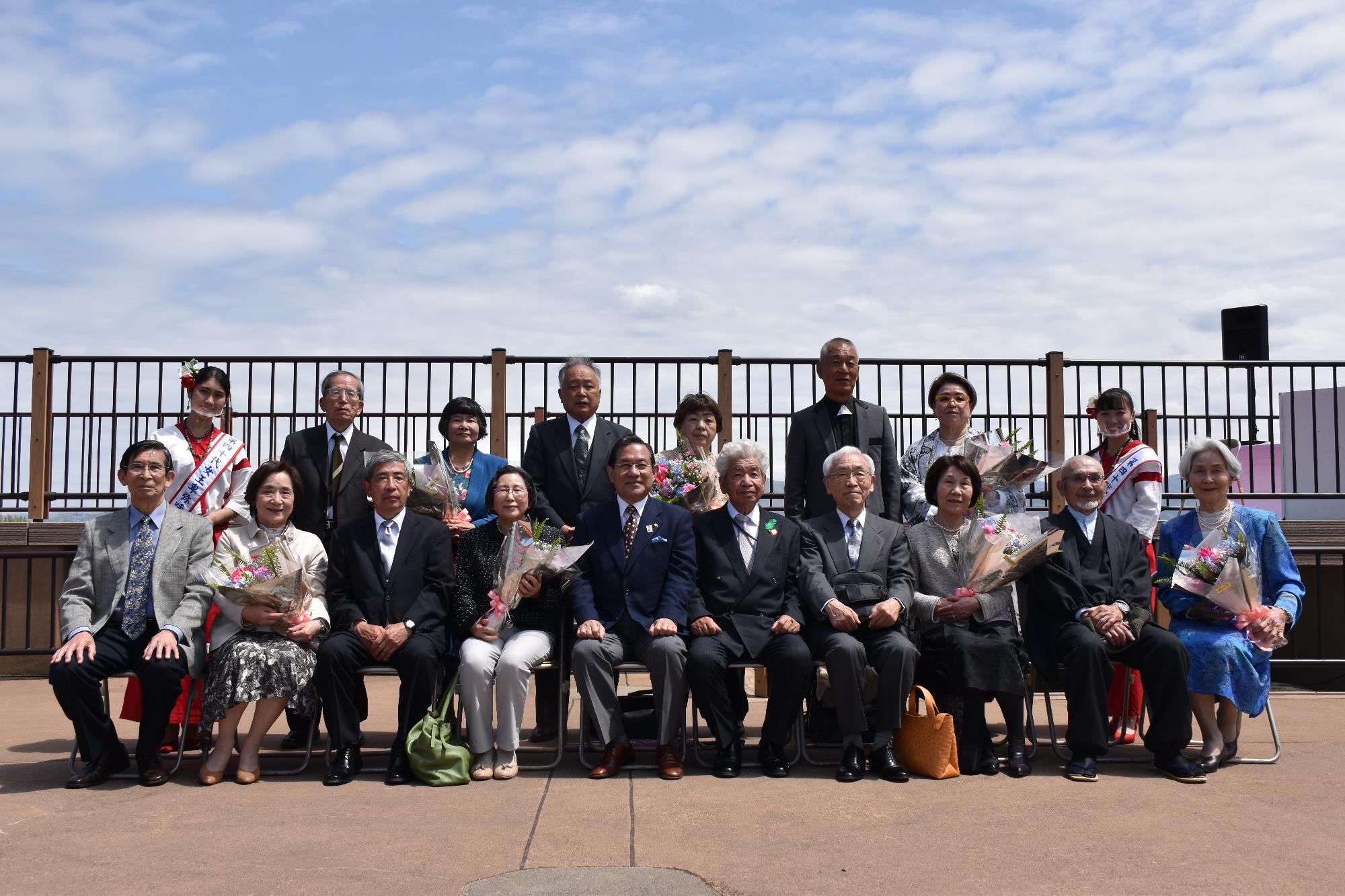 天守台展望施設で椅子に並んで座り記念撮影をする金婚式参加者夫婦7組と市長、飯田会長女王卑弥呼の二人
