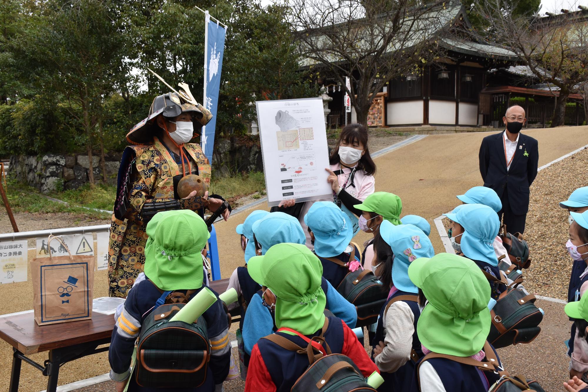 大砲の玉を持って説明をする鎧兜を付けた武将と、熱心に聞く幼稚園児たち