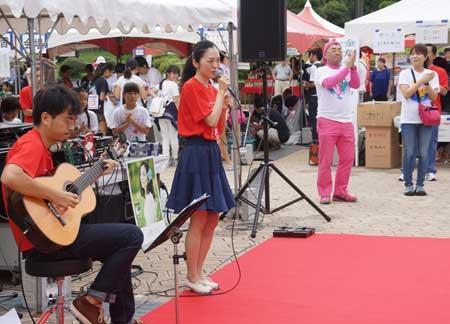 大会の会場の外にある赤いステージの上で赤いTシャツ姿の男性のアコースティックギターの演奏に合わせてマイクで歌う同じ赤いTシャツを着ている女性の写真