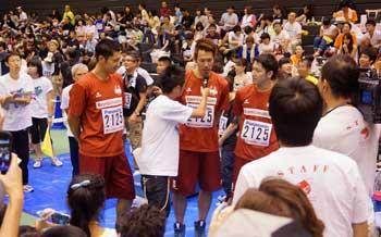 会場内で白いTシャツ姿の男性にマイクを顔に近づけられる赤いTシャツにショートパンツ姿の男性と男性のそばに立つ男性と同じ服装の2人の男性の写真