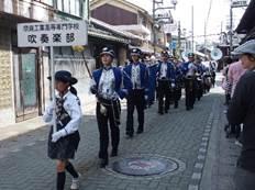 プラカードを持った女子生徒を先頭に、楽器を持った青い衣装を着た学生が街道をパレードする写真