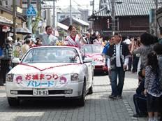 「お城まつりパレード」と書かれた横断幕が前部に貼られた白いオープンカーに乗った、着物を着た女性と白いスーツの男性がパレードしている処の写真