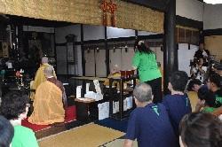 緑色のTシャツを着た女性の前に僧侶が座っているお寺の本堂の写真