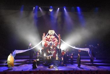 青いライトが当てられた赤と白の舞台美術のセットの下で歌劇を披露している写真
