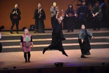 中央で両手を上げてジャンプする人と、ピンクやグレーやブラックの和服を着た人々が舞台で歌い踊っている写真