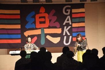 ホールで楽（GAKU)と書かれた幕の前で演奏している2人組と聞いている聴衆たちの写真