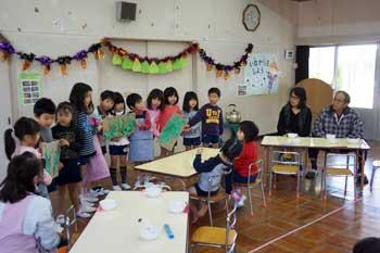 半円形に並べられた机を囲んで立って歌を歌っている幼稚園児の写真
