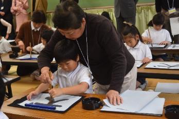 テーブルの上で書道をしている子供と一緒に筆をもって指導している女性の写真