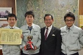 壁の前で市長と並んで賞状とトロフィーを持っている3人の男性の写真