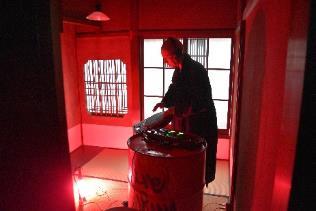 赤く照らされた和室でドラム缶の上の楽器を使って演奏をする和服姿の男性の写真