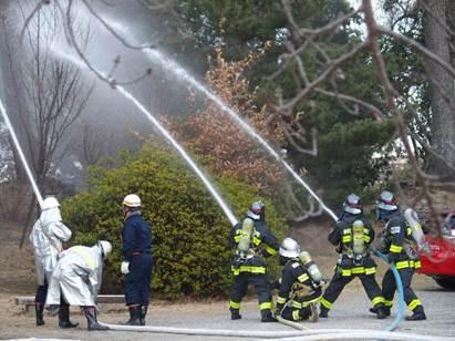 郡山城跡の敷地内で上空に向かって3本の消防用のホースを使って放水する消防服姿の7人の写真