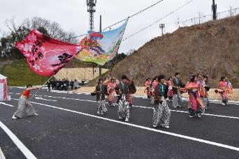 道路上でピンク色の大きな桜の花と建物の絵が描かれた大きな旗をそれぞれ持つ2人の人物と2人の前方でカラフルな衣装を着用した男女たちの写真