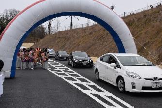 道路に設置された白い空気で膨らませたアーチをくぐり抜けるように走行する4台の自動車を見つめるカラフルな衣装を着た人々の写真