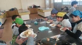 教室内の調理台で白い皿に盛りつけられた玉ねぎと鶏肉を使ったフィリピンの料理を食べている女性や男女の生徒たちの写真