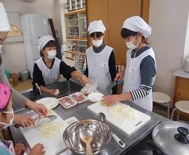 教室内の調理台の上でまた板の上の玉ねぎを包丁で切っているマスクにかっぽう着姿の女性と生徒などフィリピンの料理を作っている生徒たちの写真