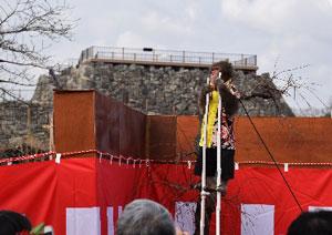 遠くに見える城の土台を背景に紅白幕のそばで白い竹馬に乗っている法被を着た猿と見つめる来場者たちの写真